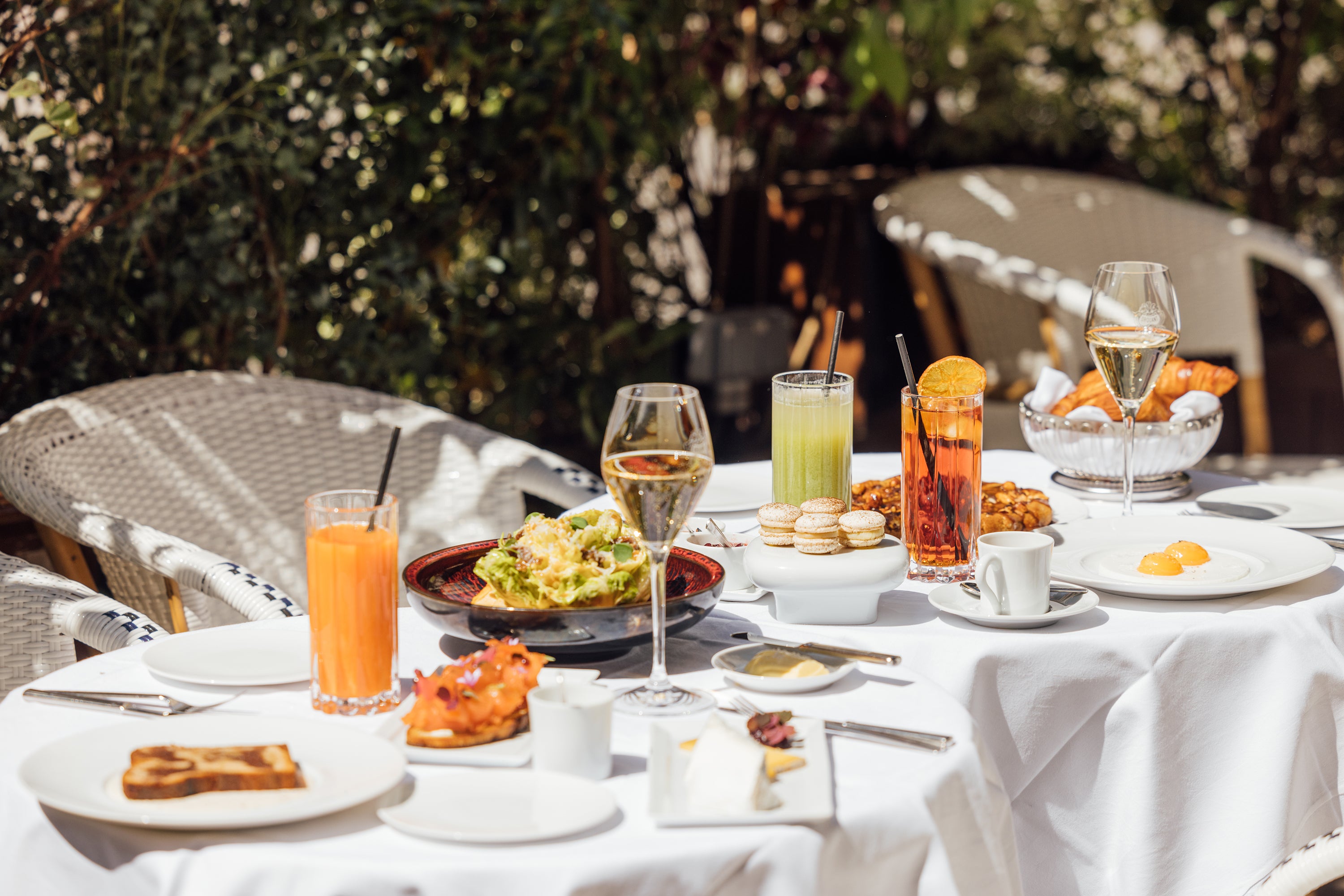 A outdoor brunch table featuring various drinks, a salad, pastries, and desserts against a green backdrop.
