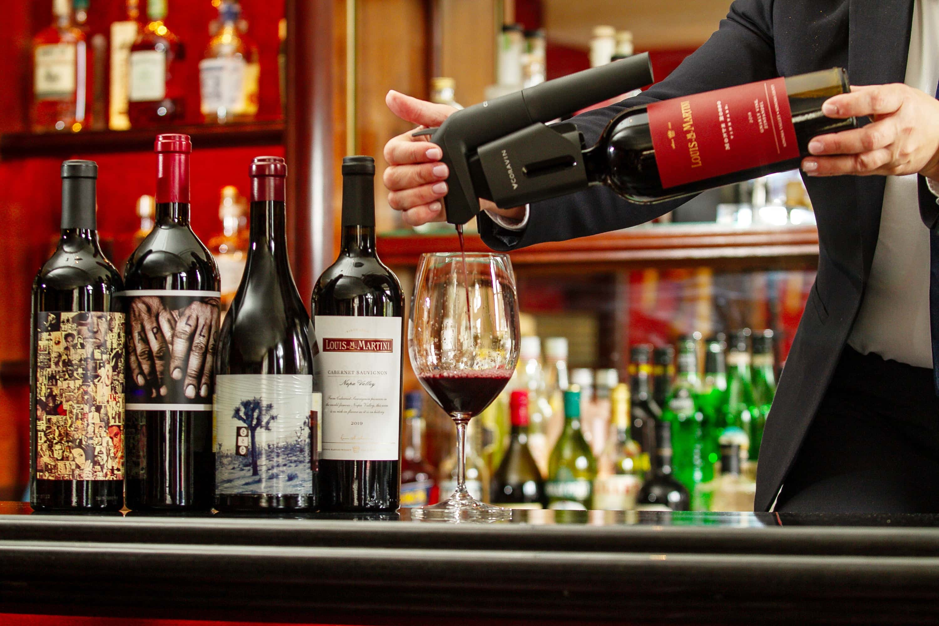 A waiter pours red wine from a bottle into a glass, alongside various wine bottles on the bar.