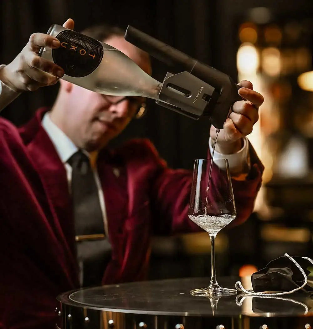 Bartender in a burgundy suit pouring liquid from a frosted bottle into a clear glass on a round table.