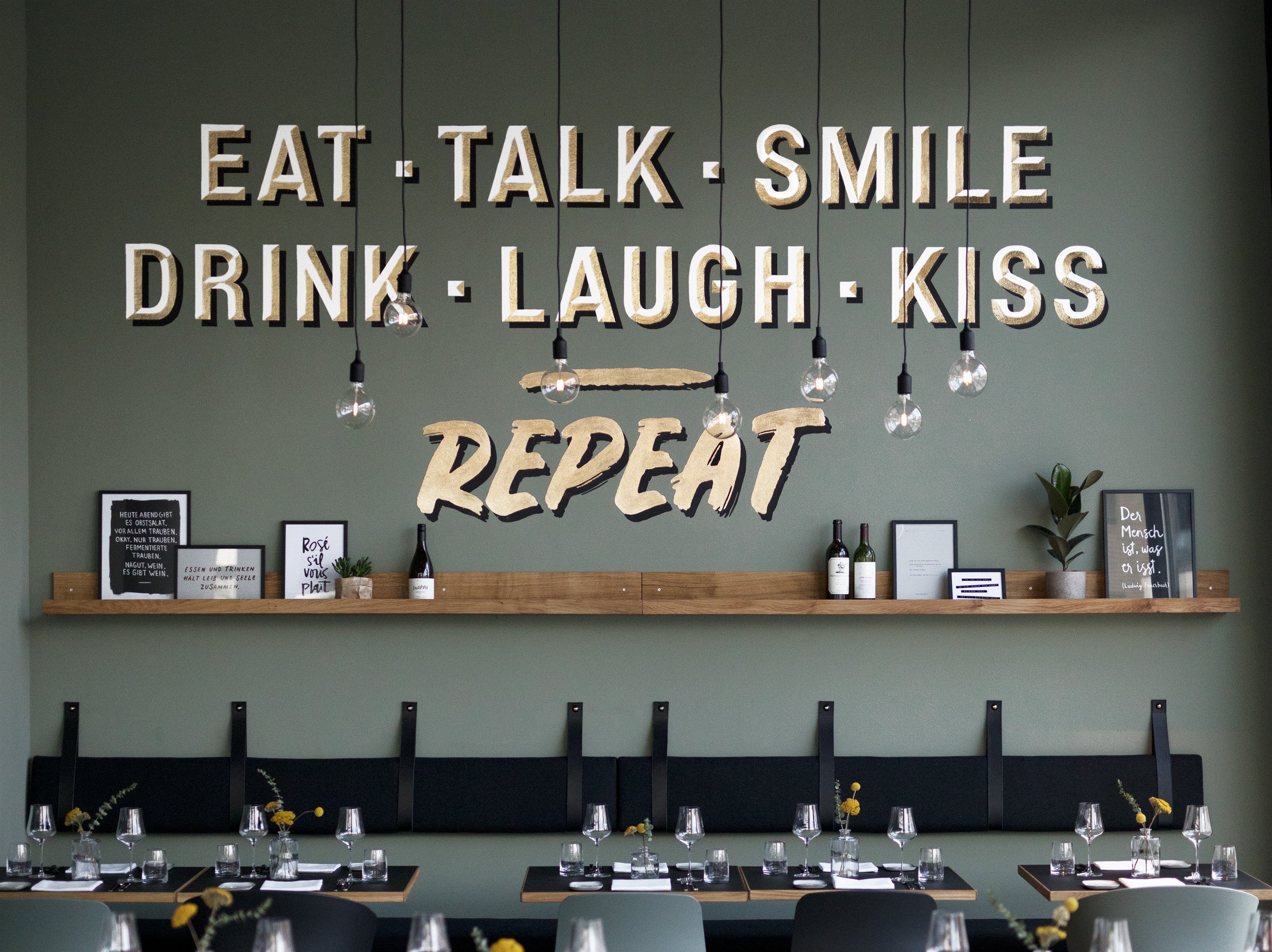 Wall decor featuring the phrases "EAT, TALK, SMILE, DRINK, LAUGH, KISS, REPEAT" with a wooden shelf displaying bottles and art.