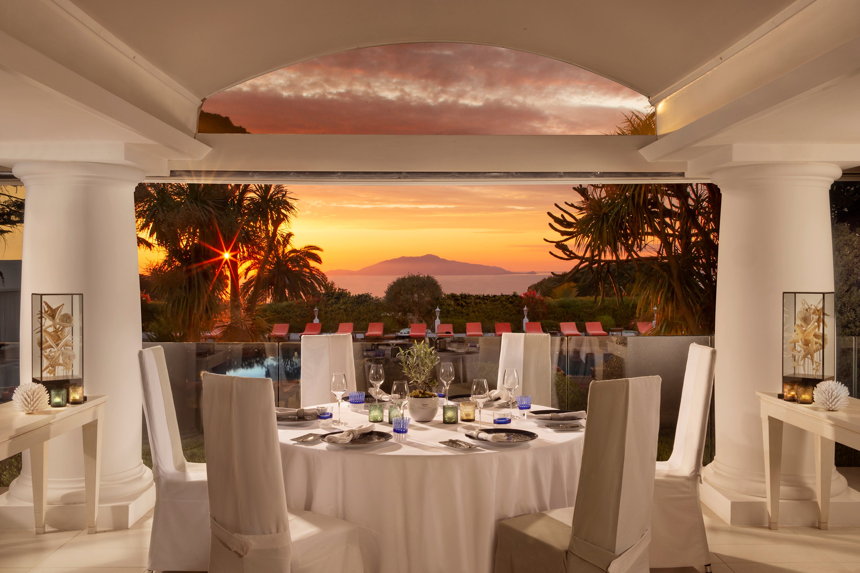 Elegant dining setup with a sunset view, featuring a white tablecloth, candles, and decorative elements by palm trees.