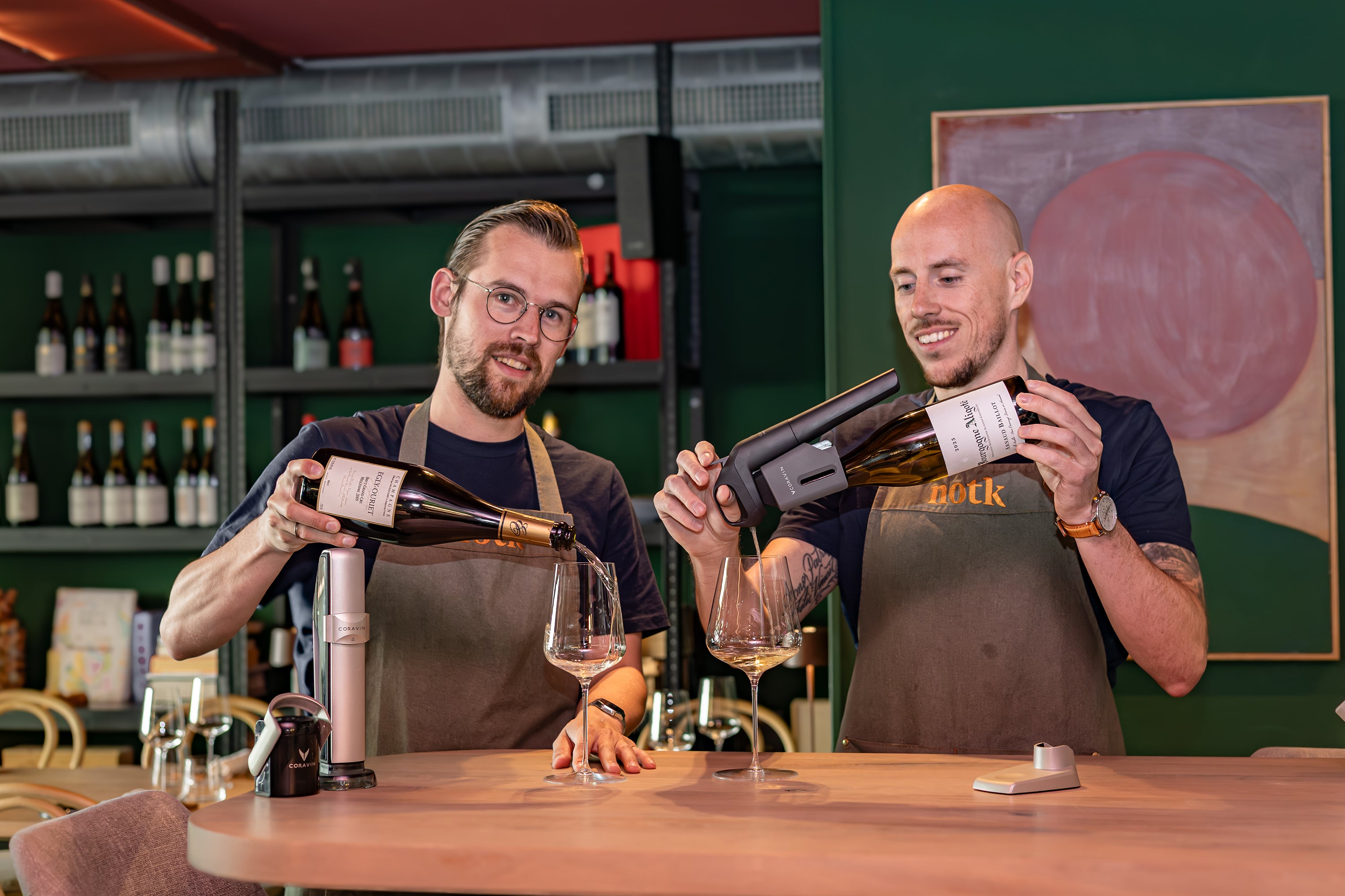 Two men in aprons pour wine into glasses at a bar, showcasing a modern wine dispensing device and bottles in the background.