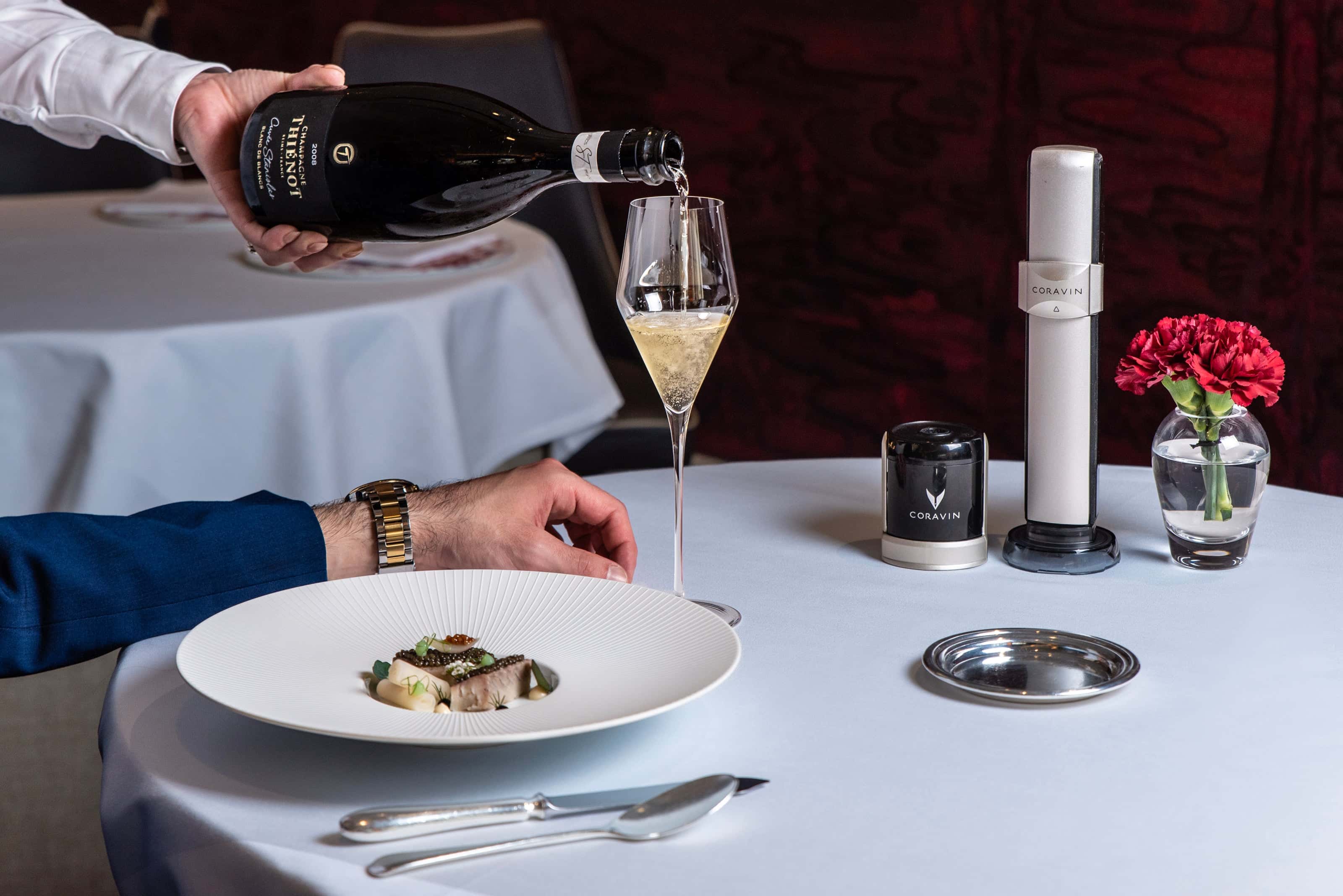 A hand pours champagne into a flute beside a gourmet dish on a table with elegant table settings and a flower vase.