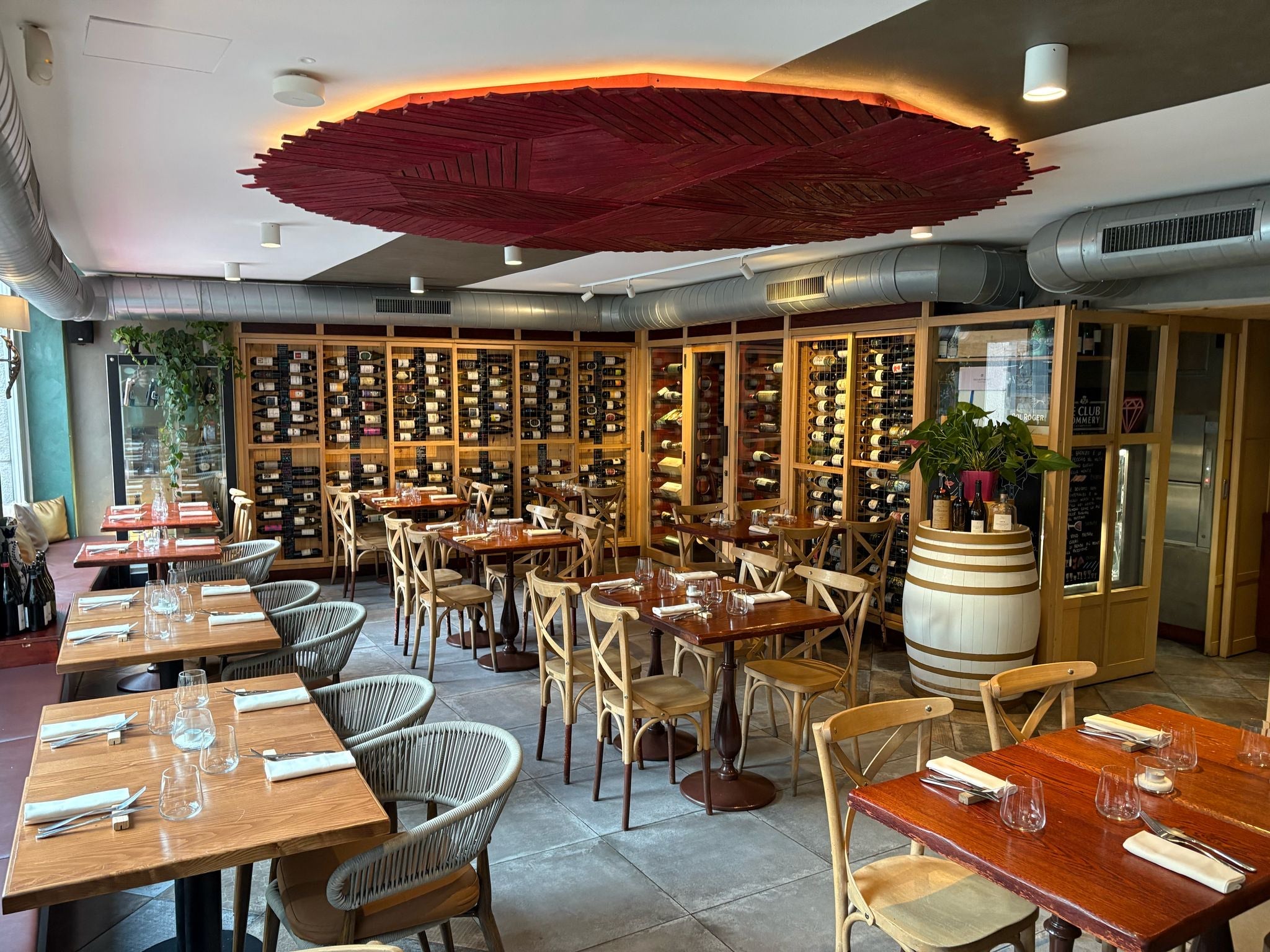 Modern restaurant interior featuring wooden tables, wicker chairs, and a wine display against the wall.