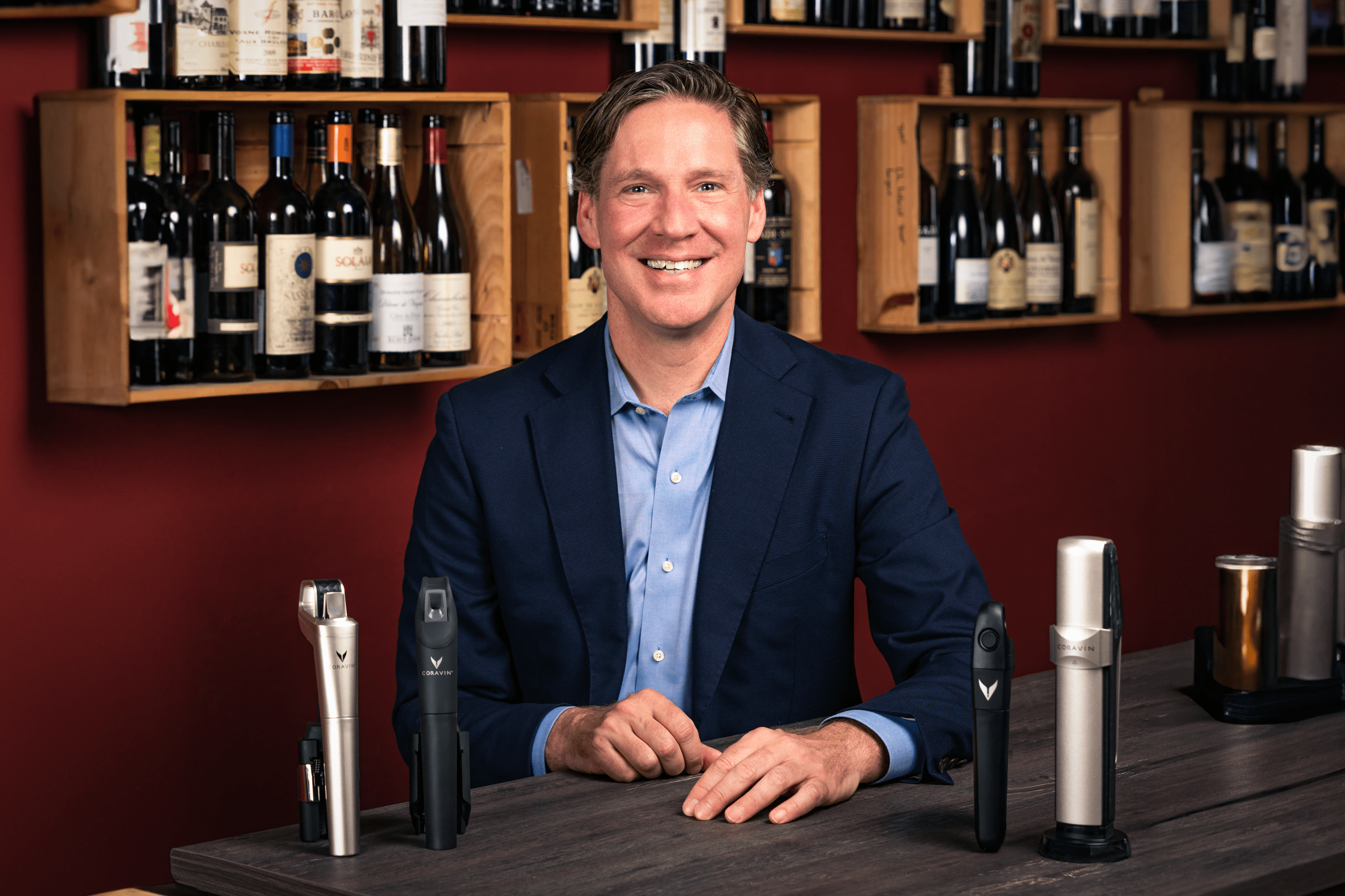 Smiling man in a blazer sits at a table with wine preservation tools and a backdrop of wine bottles on shelves.