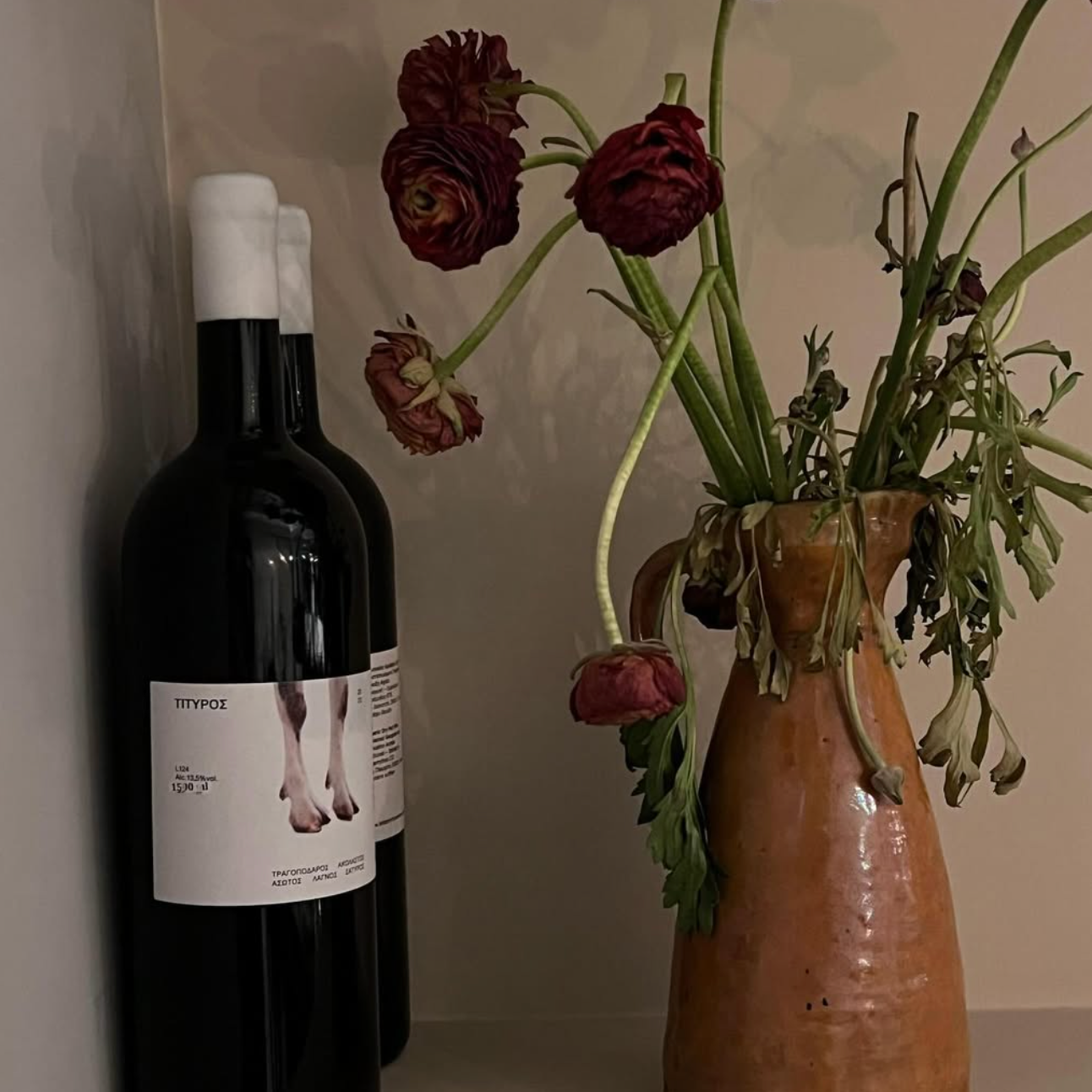 Two black wine bottles with a label featuring a photo of feet, beside a vase of wilted red flowers.
