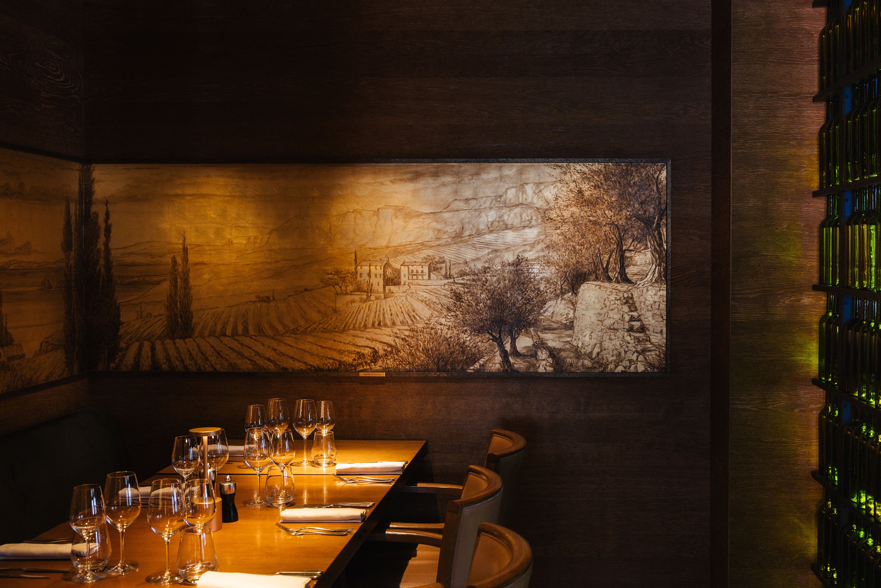 Elegant dining table set with glassware, a wooden wall featuring a vineyard landscape, and a wine display in a restaurant.