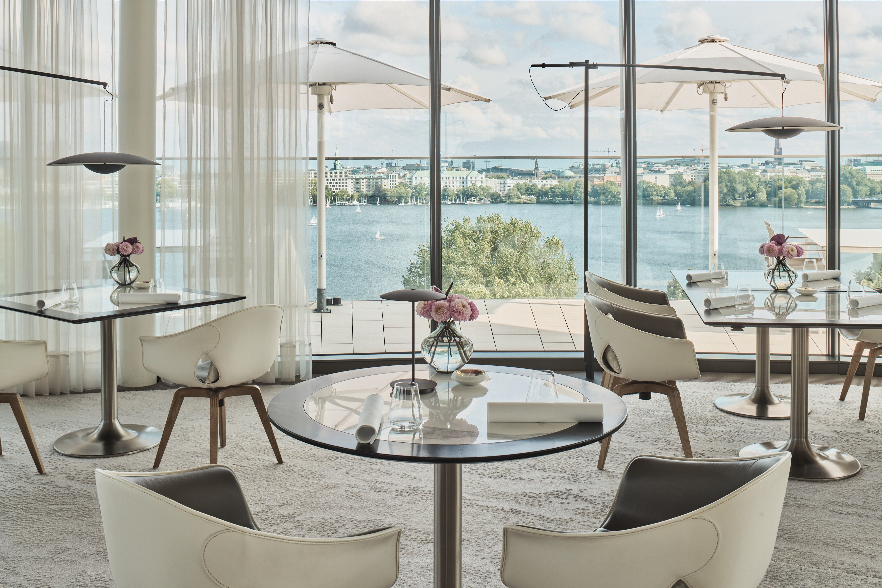 Modern dining area with glass tables, white chairs, and a scenic view of the water and city through large windows.