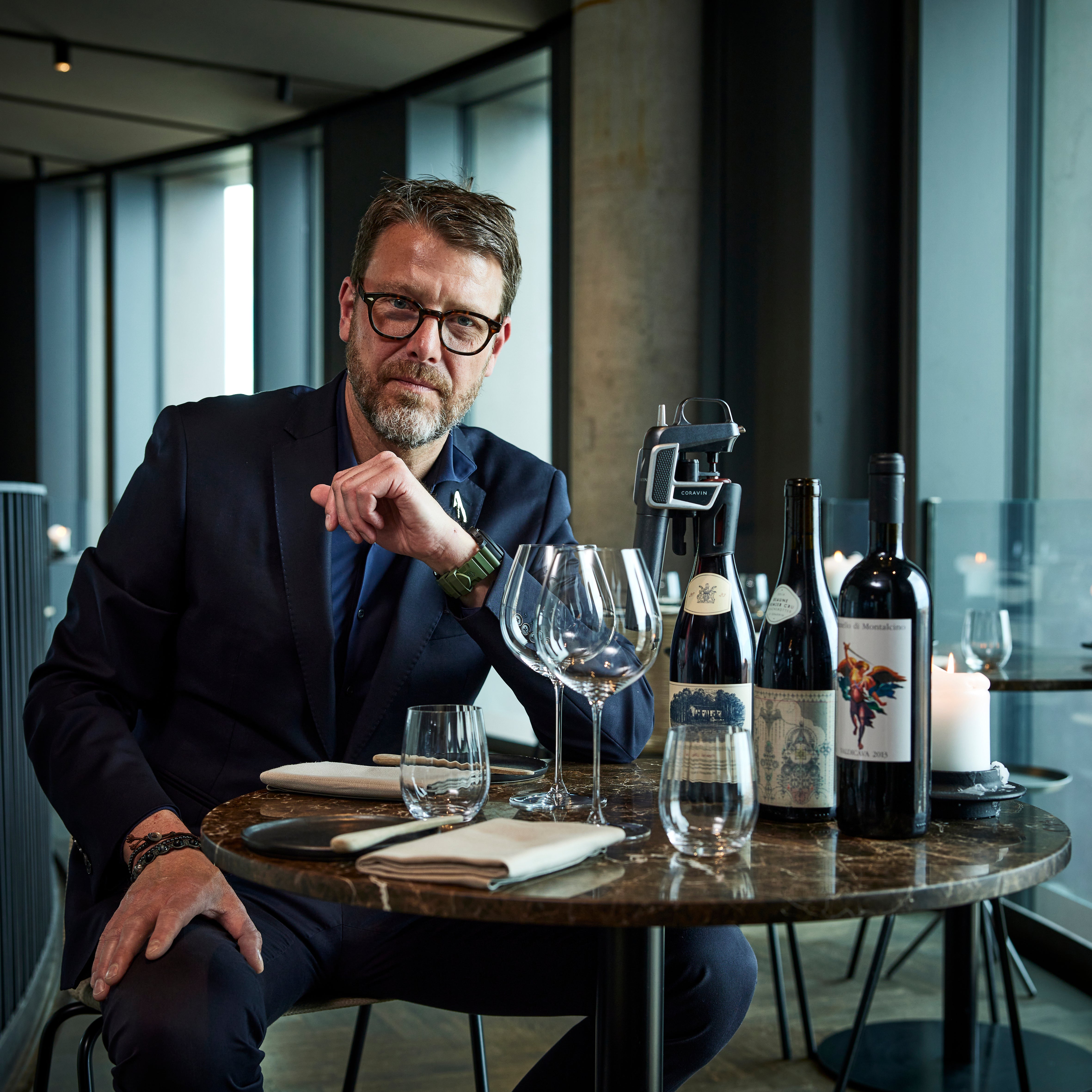 A man in a suit sits at a table with wine bottles, glasses, and candles, looking thoughtfully at the viewer.