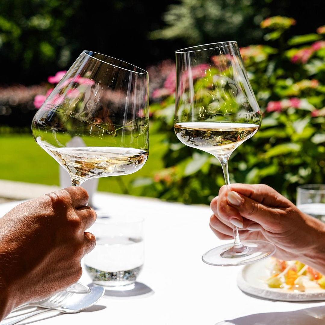 Two hands clinking white wine glasses in a garden setting, with a blurred background of colorful flowers and a table setting.