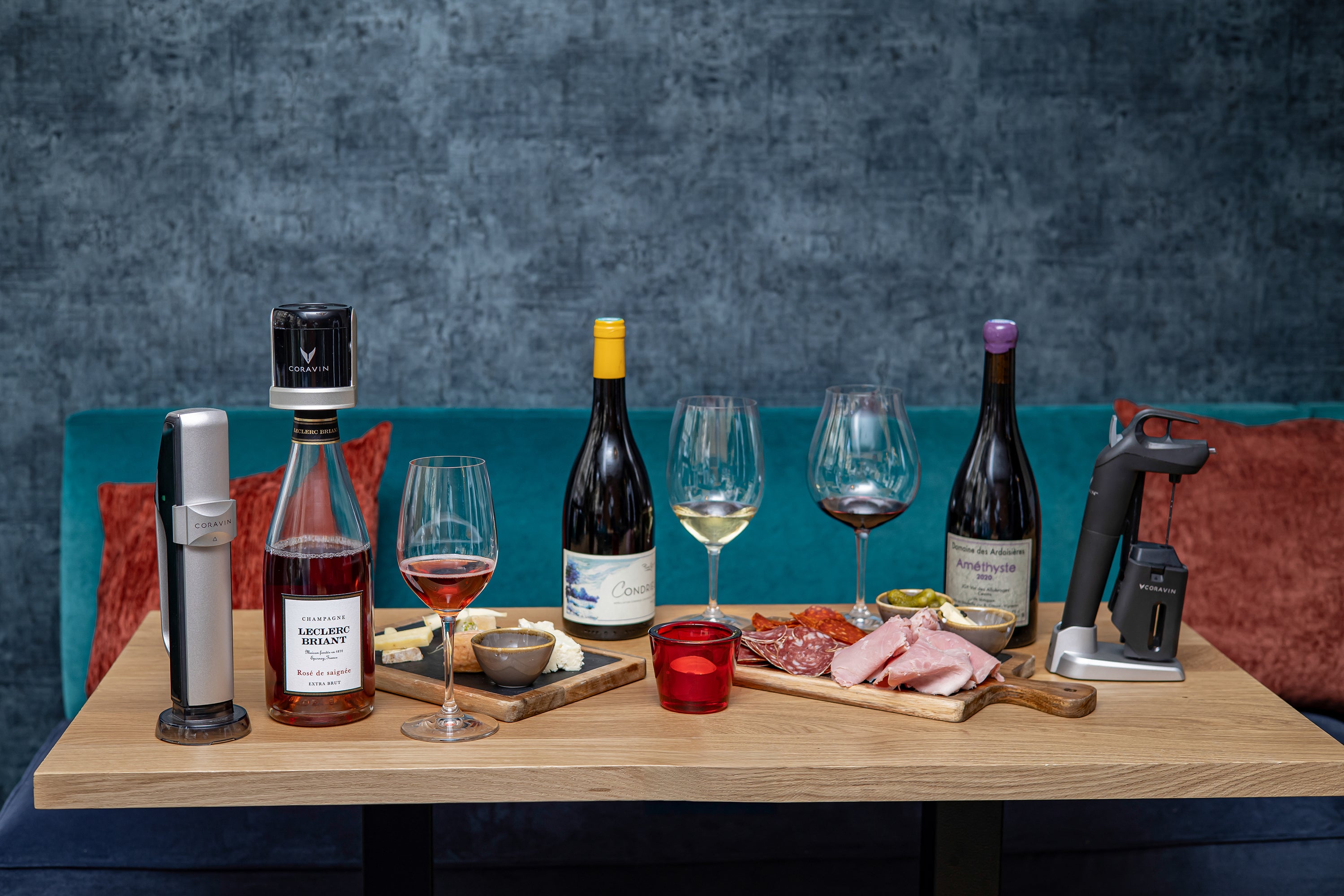Wine bottles, glasses, gourmet cheeses, meats, and a Coravin wine opener arranged on a wooden table with a blue backdrop.