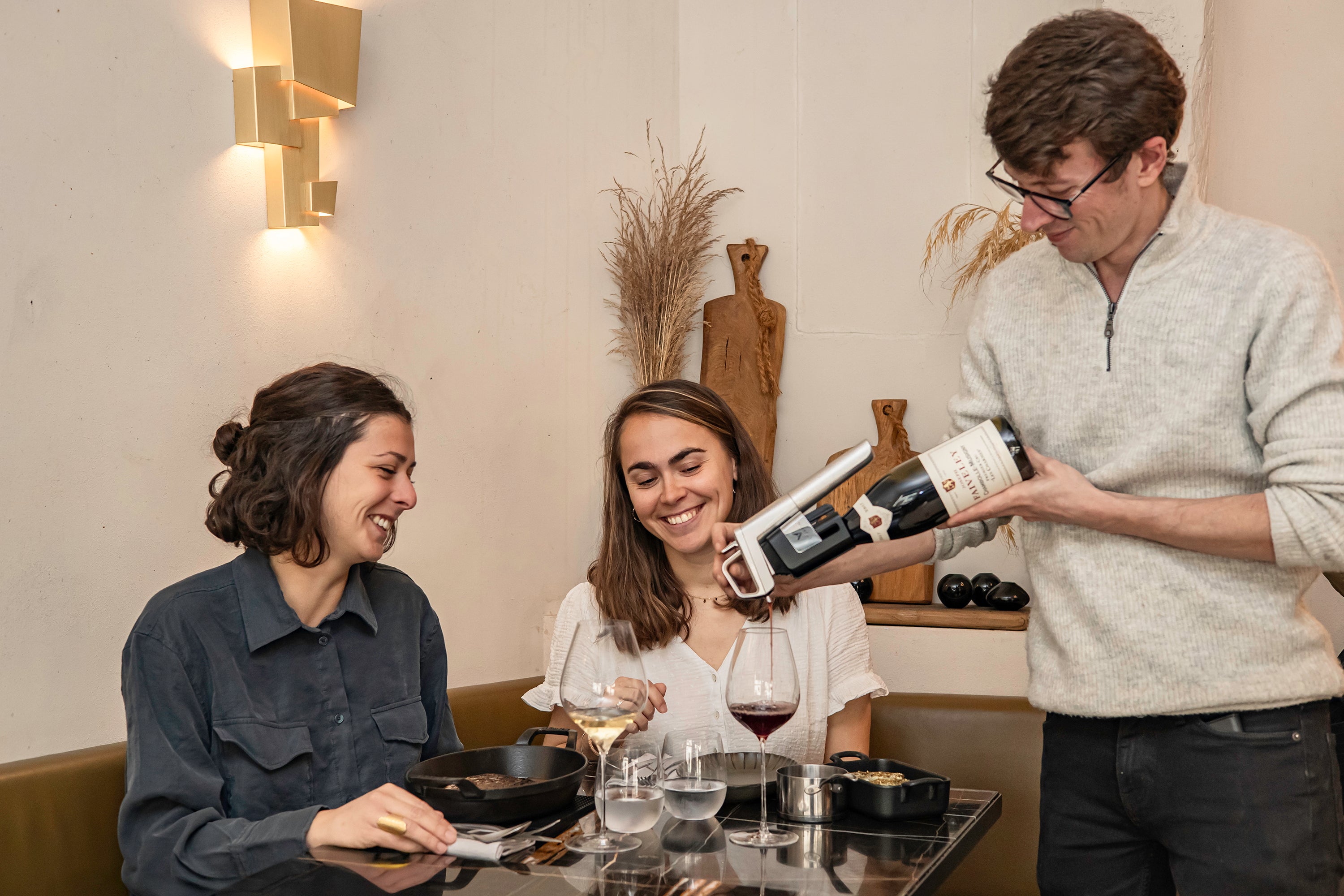 Two women enjoy wine at a table while a waiter pours red wine from a bottle, with food and glassware visible.