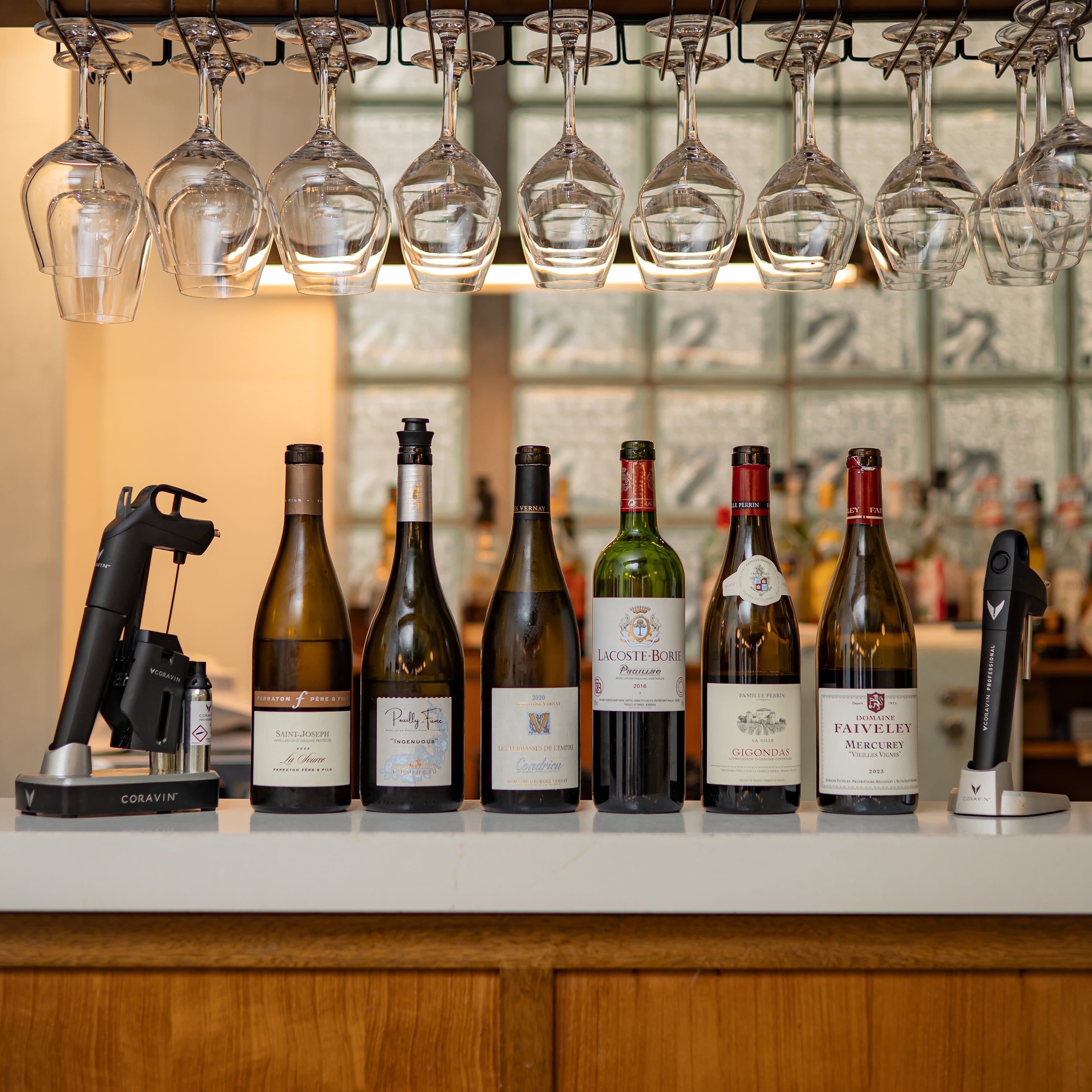 A selection of wine bottles lined up on a bar with hanging glasses above and a Coravin wine preservation tool on the left.