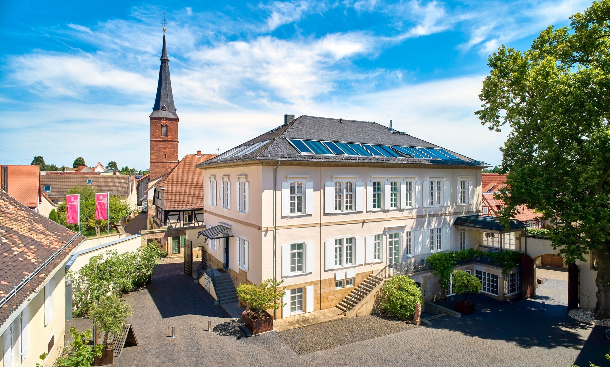 A sunny view of a historic building with solar panels, a courtyard, and a tall church steeple in the background.