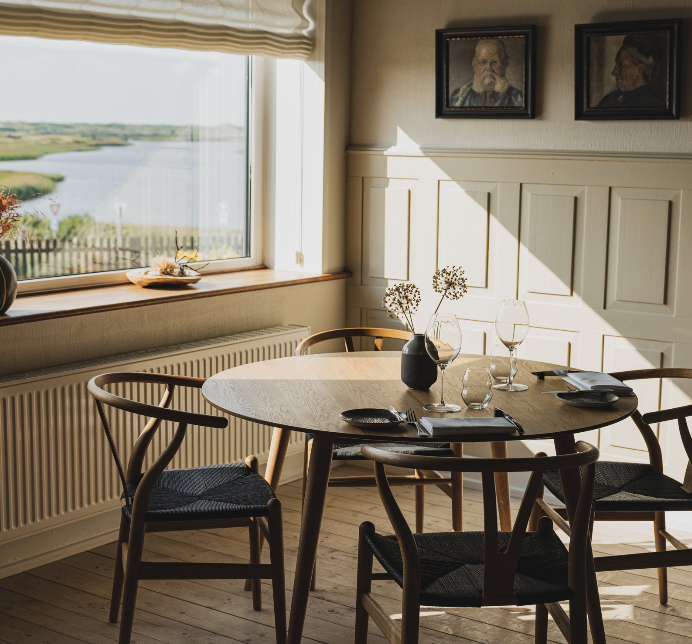 Cozy dining area with a wooden table, flower vase, and scenic window view overlooking a water body and greenery.