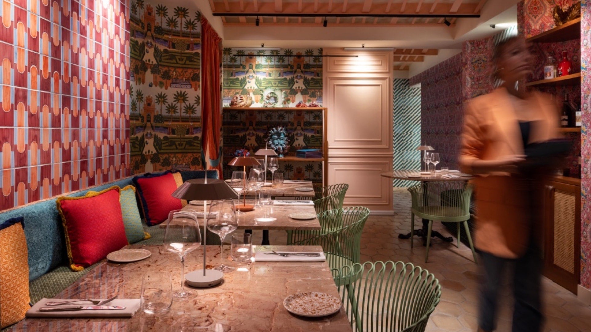 A vibrant restaurant interior featuring colorful pillows, patterned walls, and a blurred server in an orange jacket.