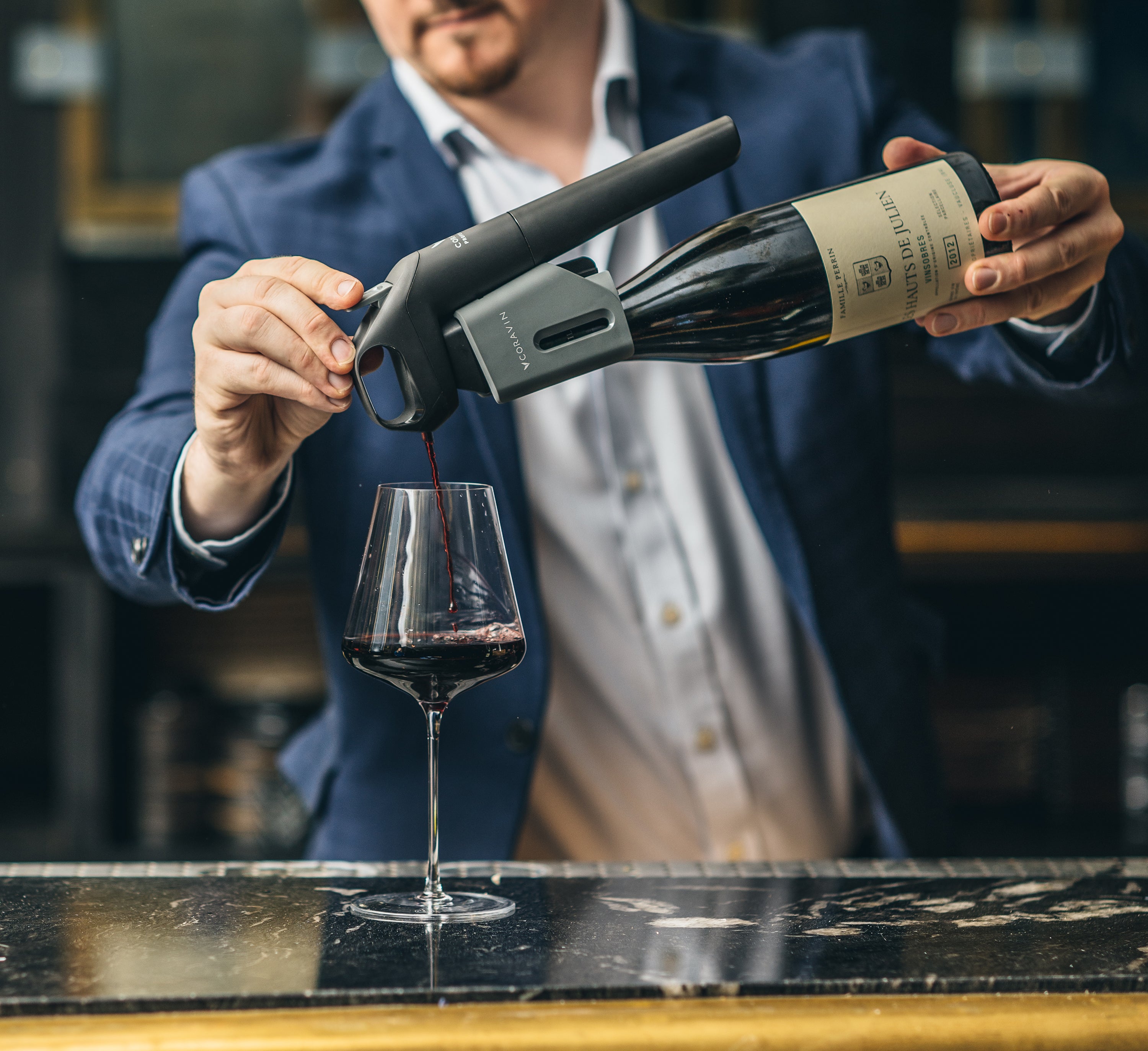 A person pours red wine from a bottle into a glass using a wine aerator, set on a polished bar countertop.