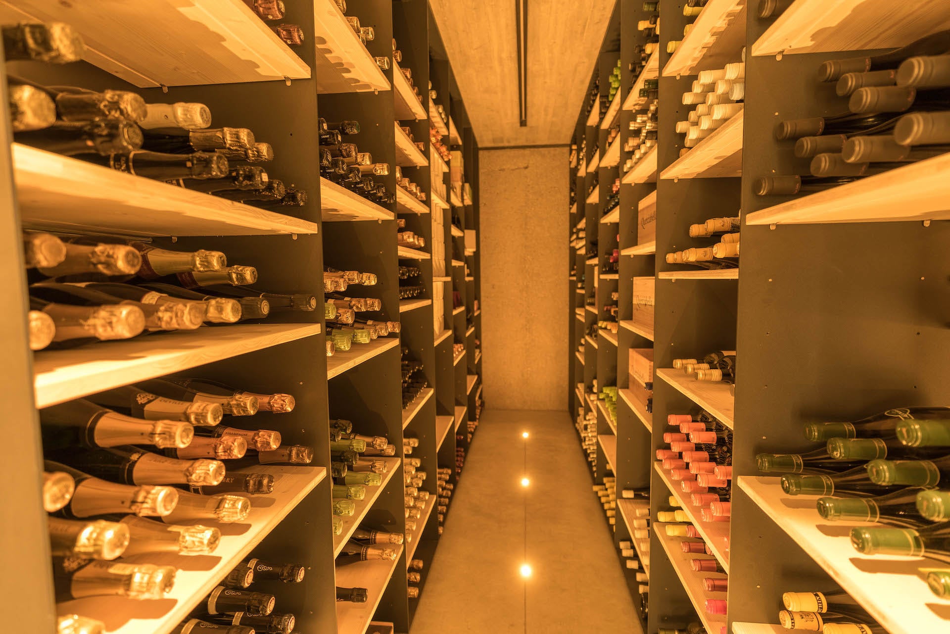 Wine storage room with wooden shelves filled with various bottles of wine, illuminated by warm lighting.