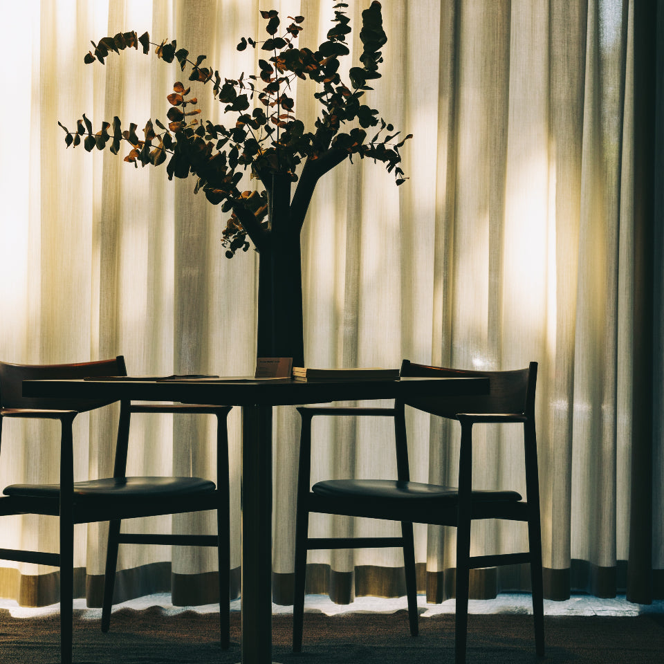 Silhouette of a vase with eucalyptus branches on a table, accompanied by two wooden chairs and sheer curtains in the background.