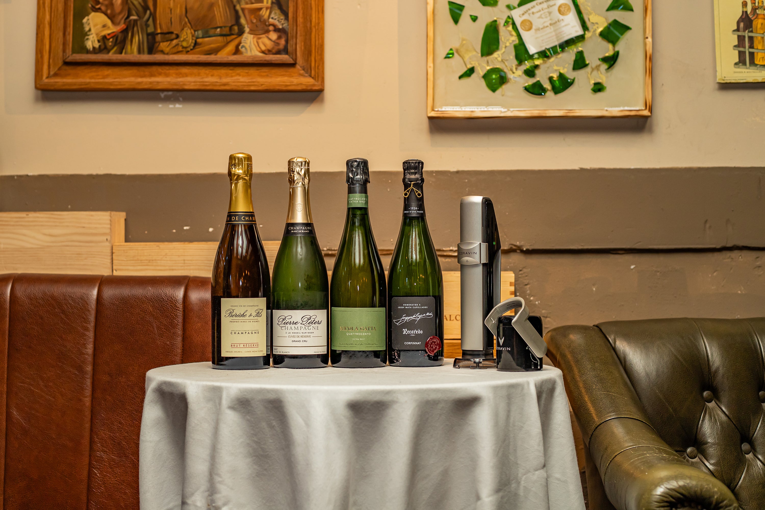 Five bottles of champagne arranged on a table, alongside a corkscrew and wine preservation tool, with a leather couch in the background.
