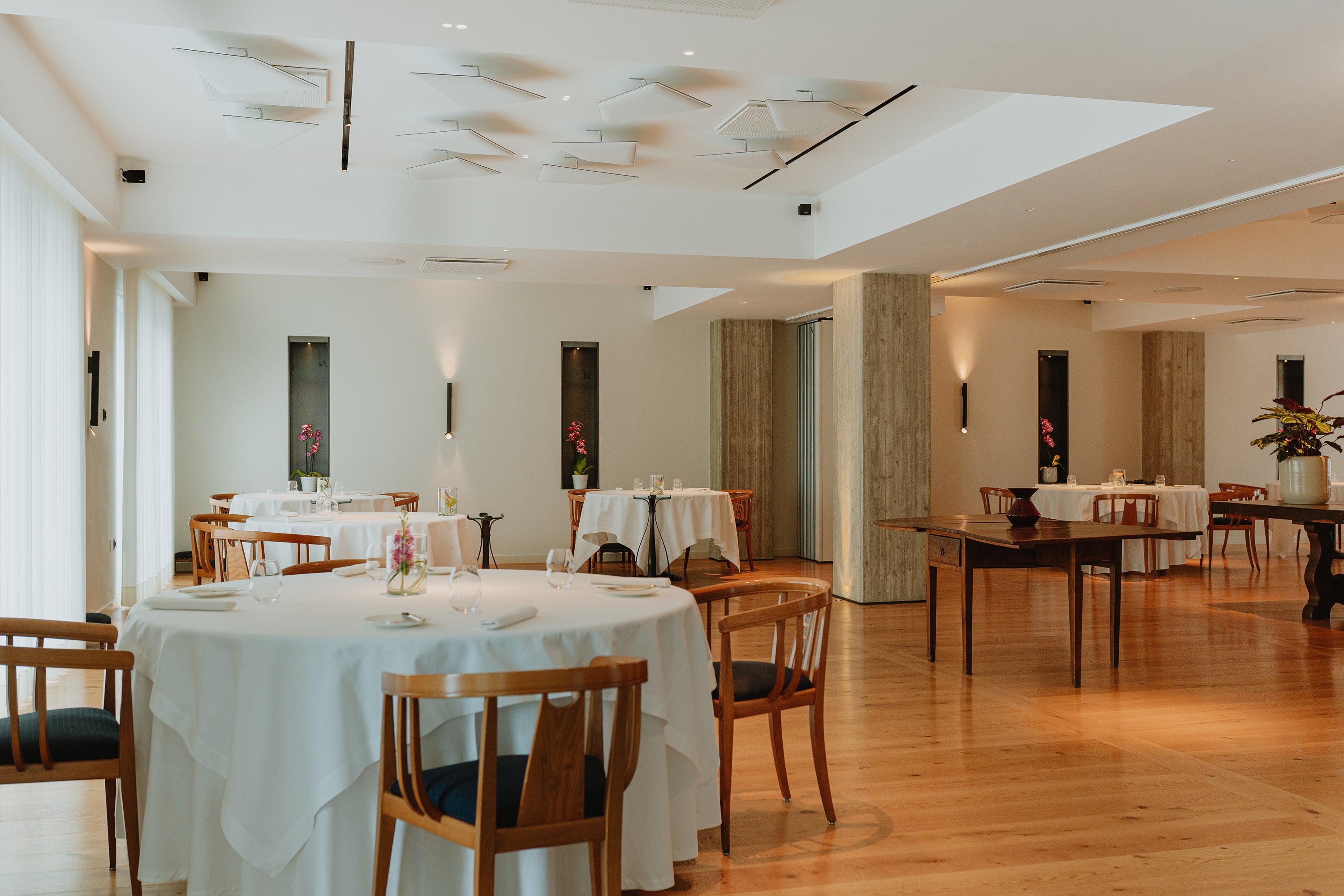 Elegant dining area with wooden chairs, round tables set with white linens, and soft lighting in a modern restaurant.