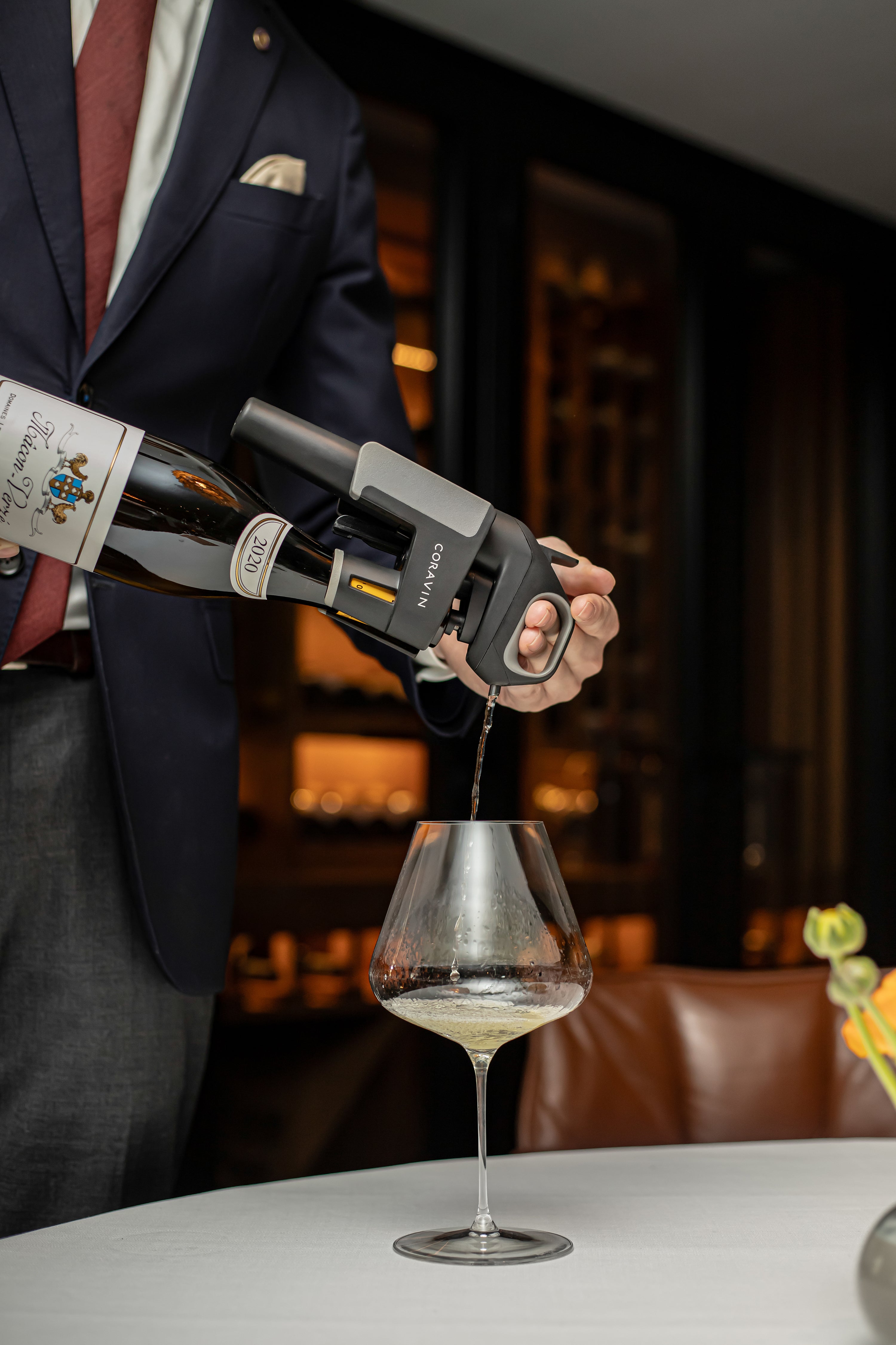 A waiter pours wine from a bottle into a large glass using a Coravin wine system at a dining table.