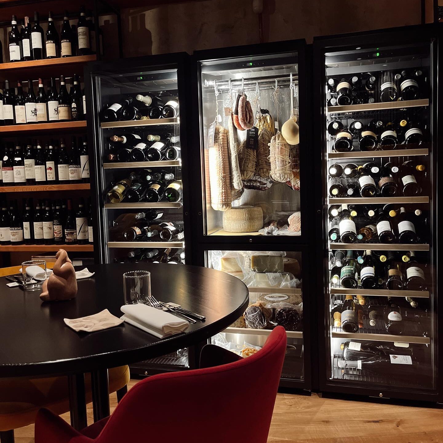 Well-stocked wine fridge with assorted wines and cured meats; wooden table set with cutlery, glass, and cloth napkin.