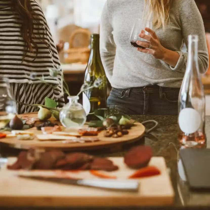 Two women standing at a kitchen counter with wine glasses, surrounded by bottles of wine and charcuterie boards filled with assorted meats, cheeses, and fruit.