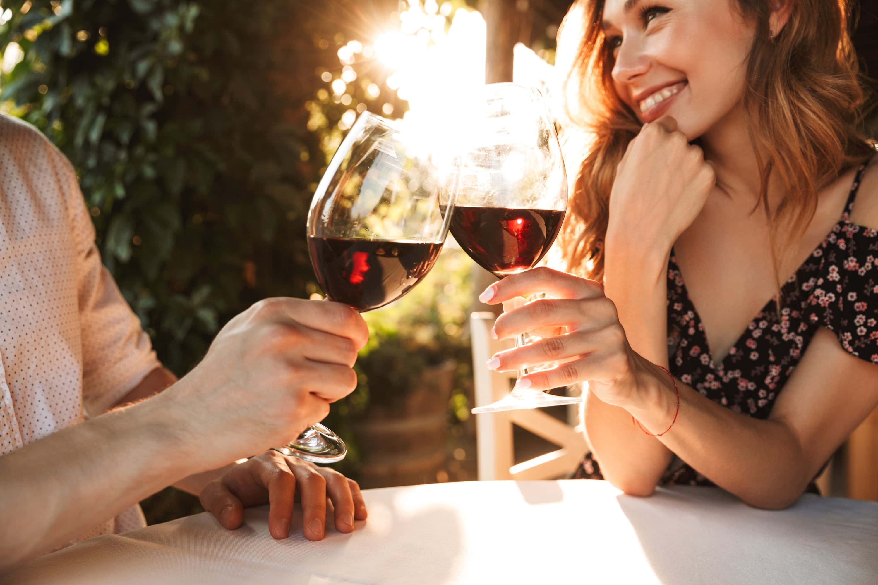 Smiling couple toasting glasses of red wine at an outdoor table during sunset, surrounded by greenery.