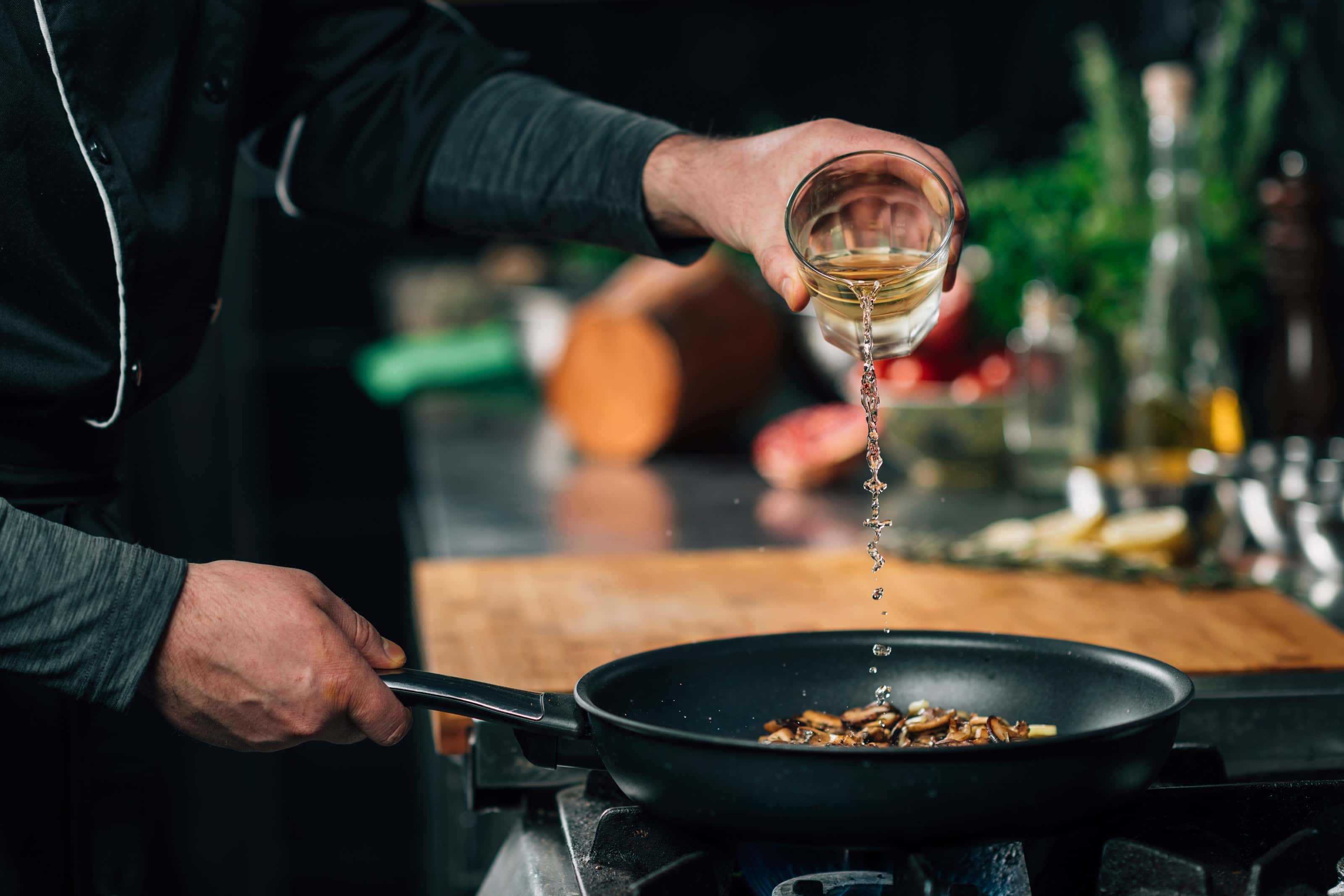 Chef pouring white wine into a skillet of mushrooms on a gas stove in a professional kitchen, with herbs and cooking ingredients in the background.