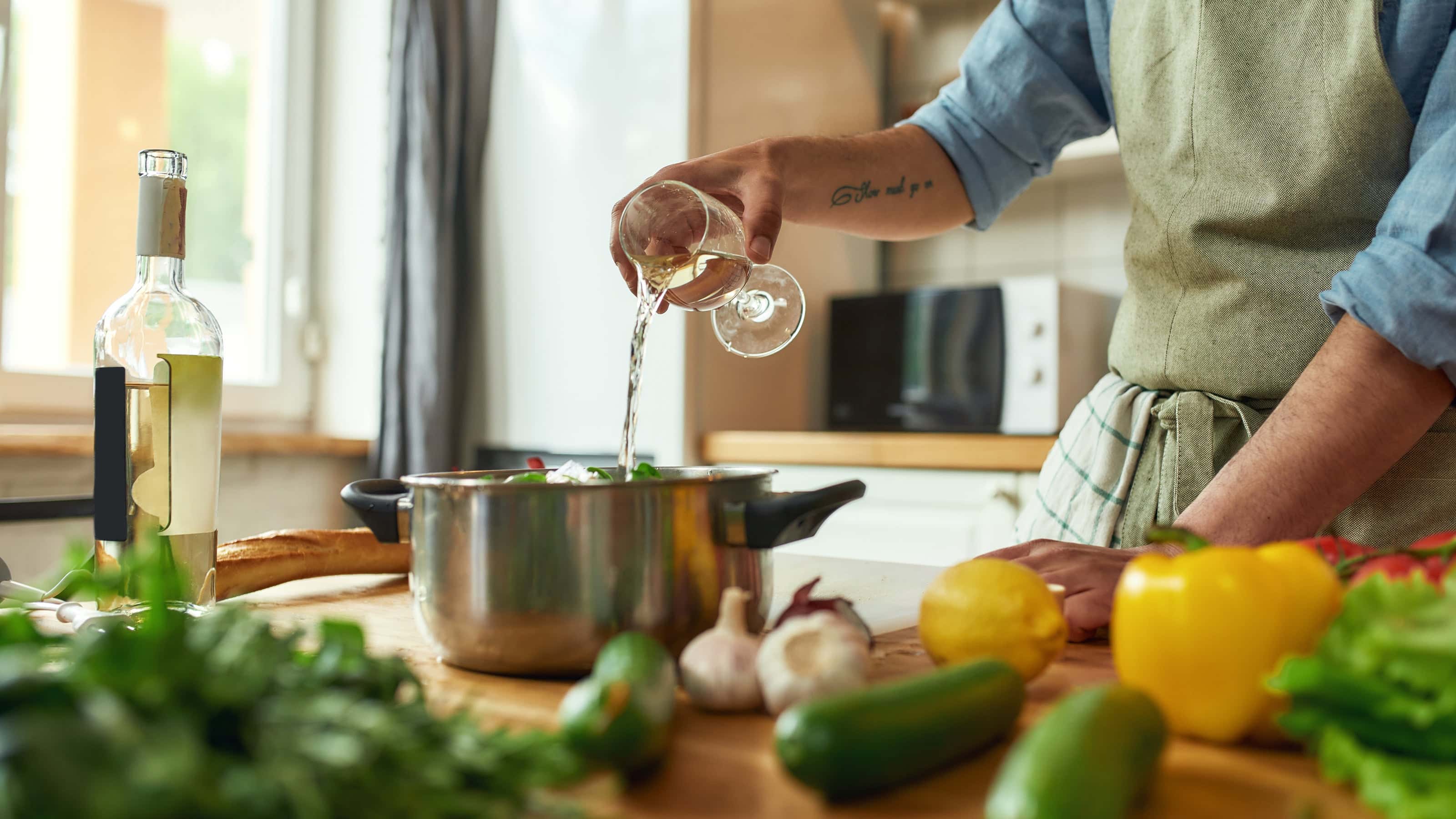 Person in apron pouring white wine from a glass into a pot on a kitchen counter, surrounded by fresh vegetables like bell peppers, zucchini, garlic, and herbs.