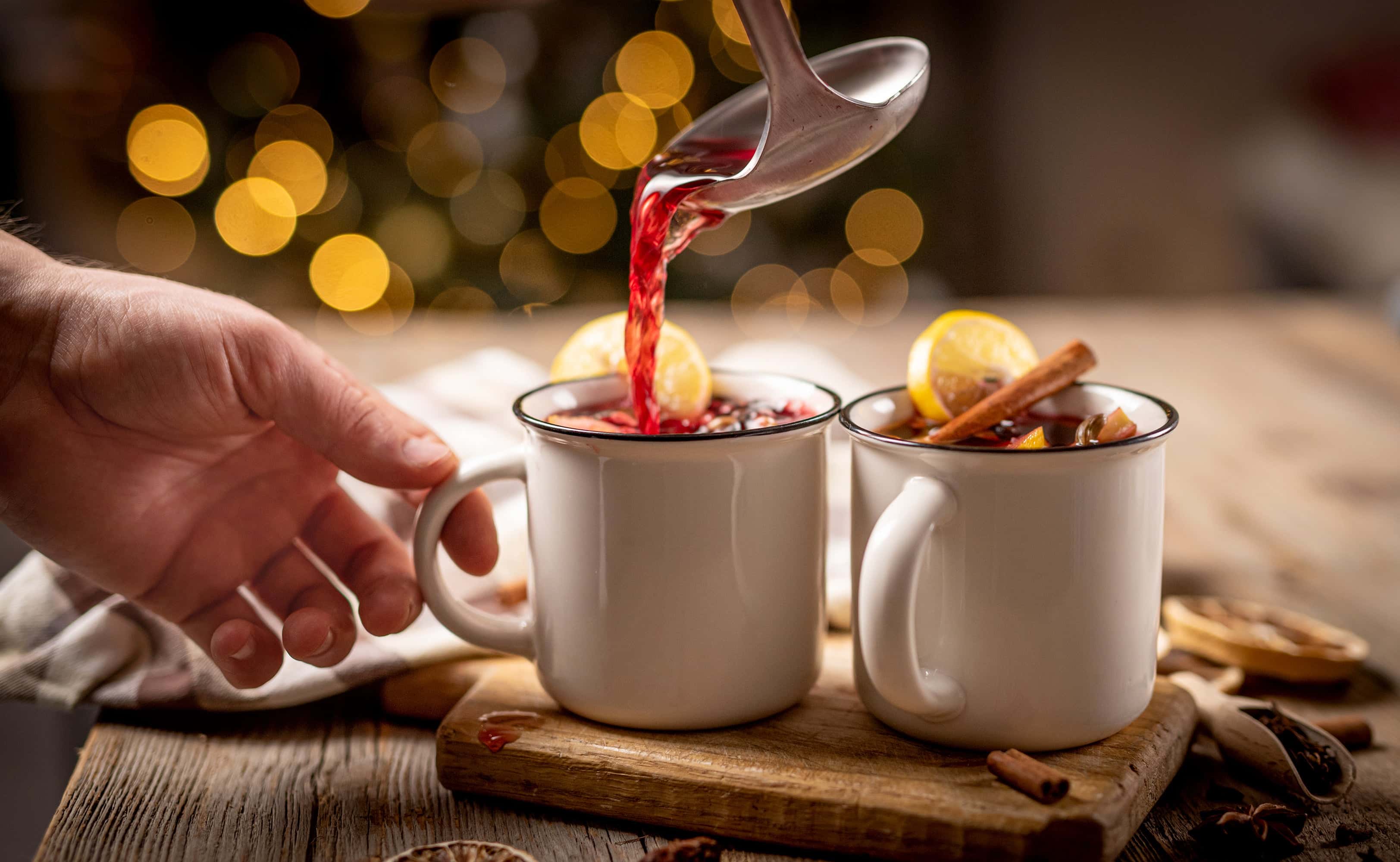 Close-up of mulled wine being poured from a ladle into white enamel mugs garnished with lemon slices and cinnamon sticks, set on a rustic wooden table with festive lights in the background.