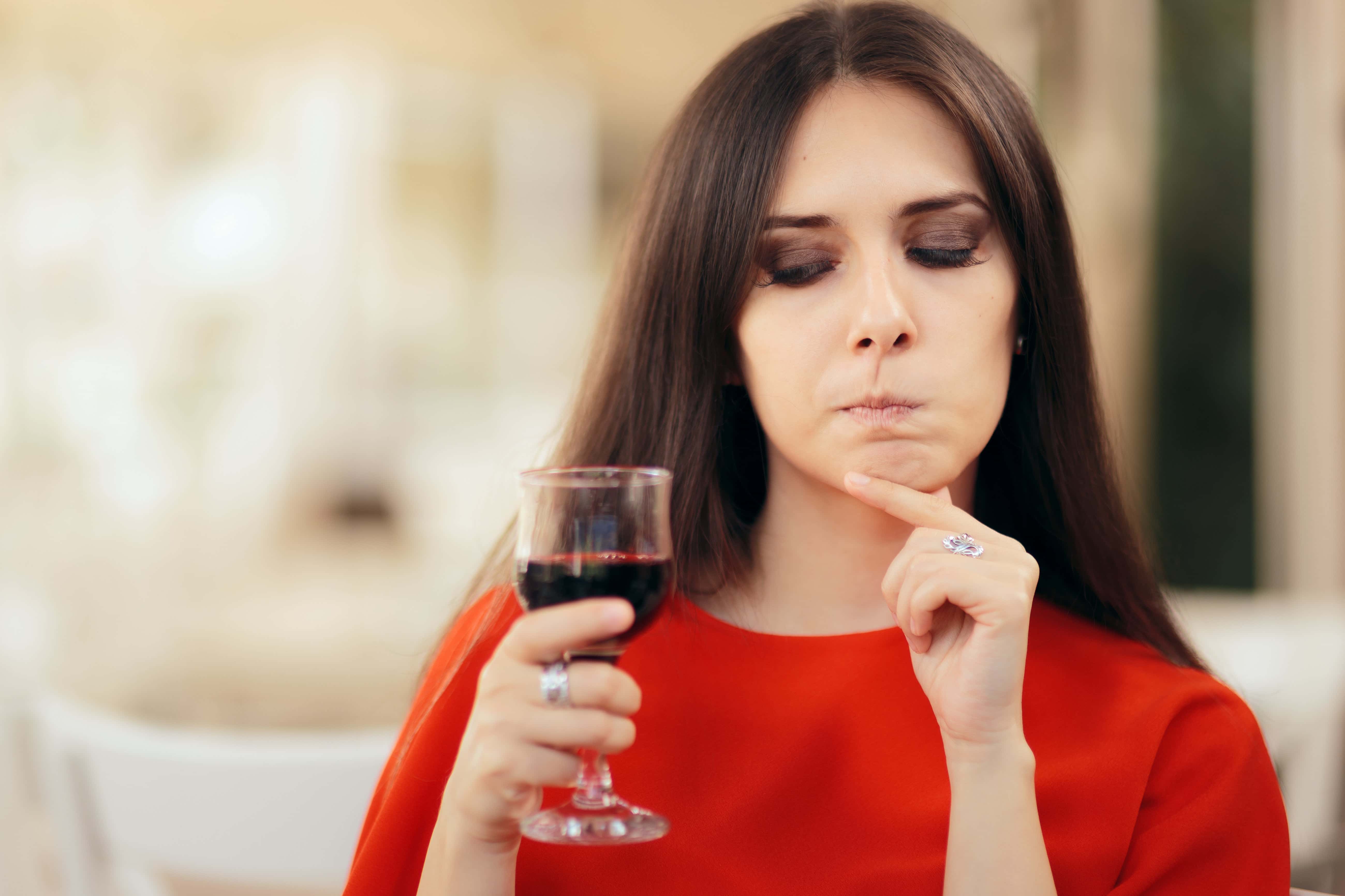 Woman in a red top thoughtfully tasting red wine from a glass, with a pensive expression and blurred background.