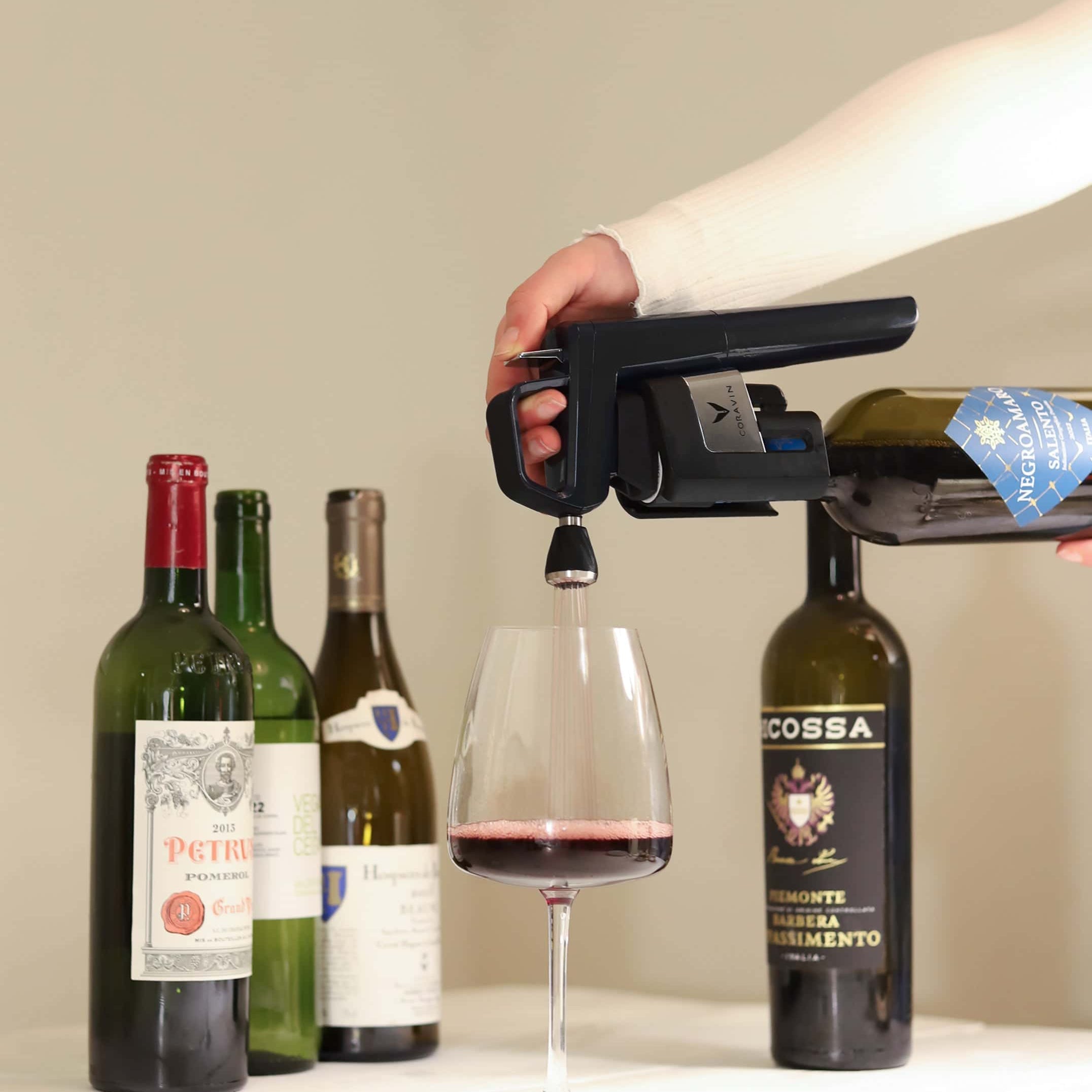 Hand using a Coravin wine preservation system to pour red wine into a glass, with several wine bottles in the background on a white tablecloth.