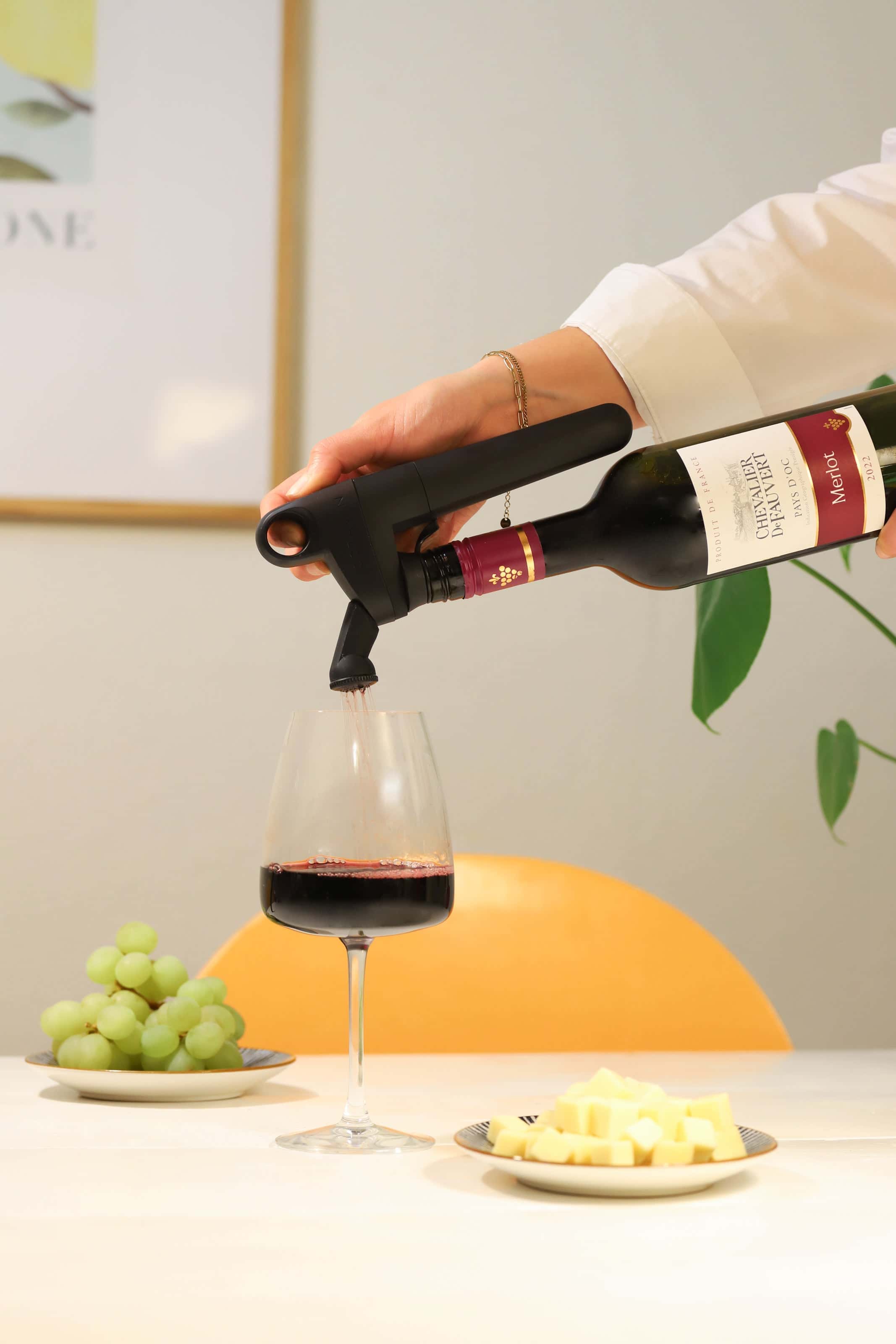 Pouring red wine from a Coravin Pivot Wine Preservation System into a glass, with grapes and cheese on a table, showcasing wine aeration and preservation.