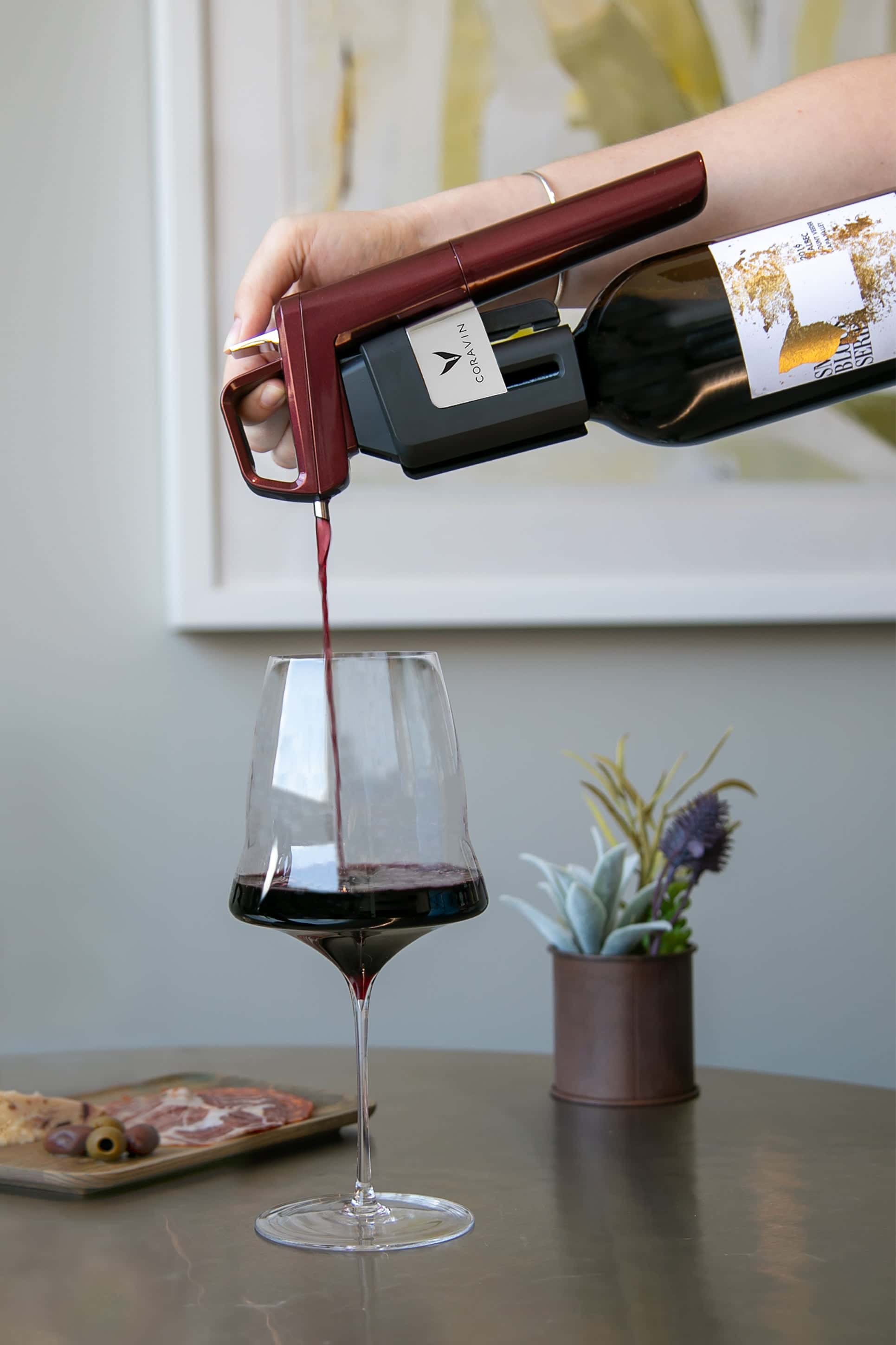 Red wine being poured into a glass using a Coravin wine preservation system, with a charcuterie board and small potted plant in the background.