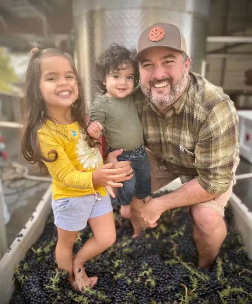 Smiling man with two young children standing barefoot in a large container filled with harvested grapes during winemaking.