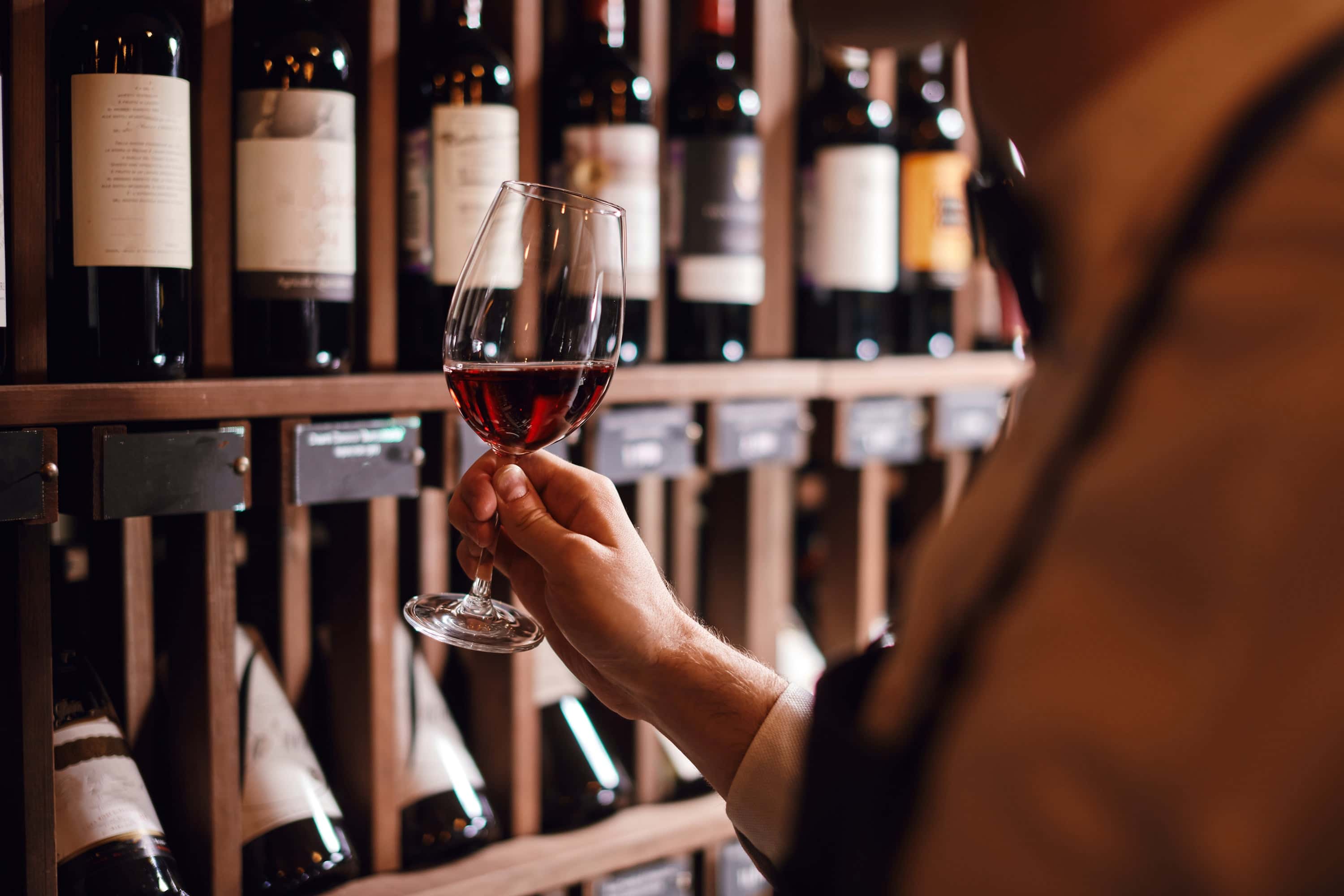 A hand holding a glass of red wine in front of a wooden wine rack filled with bottles.