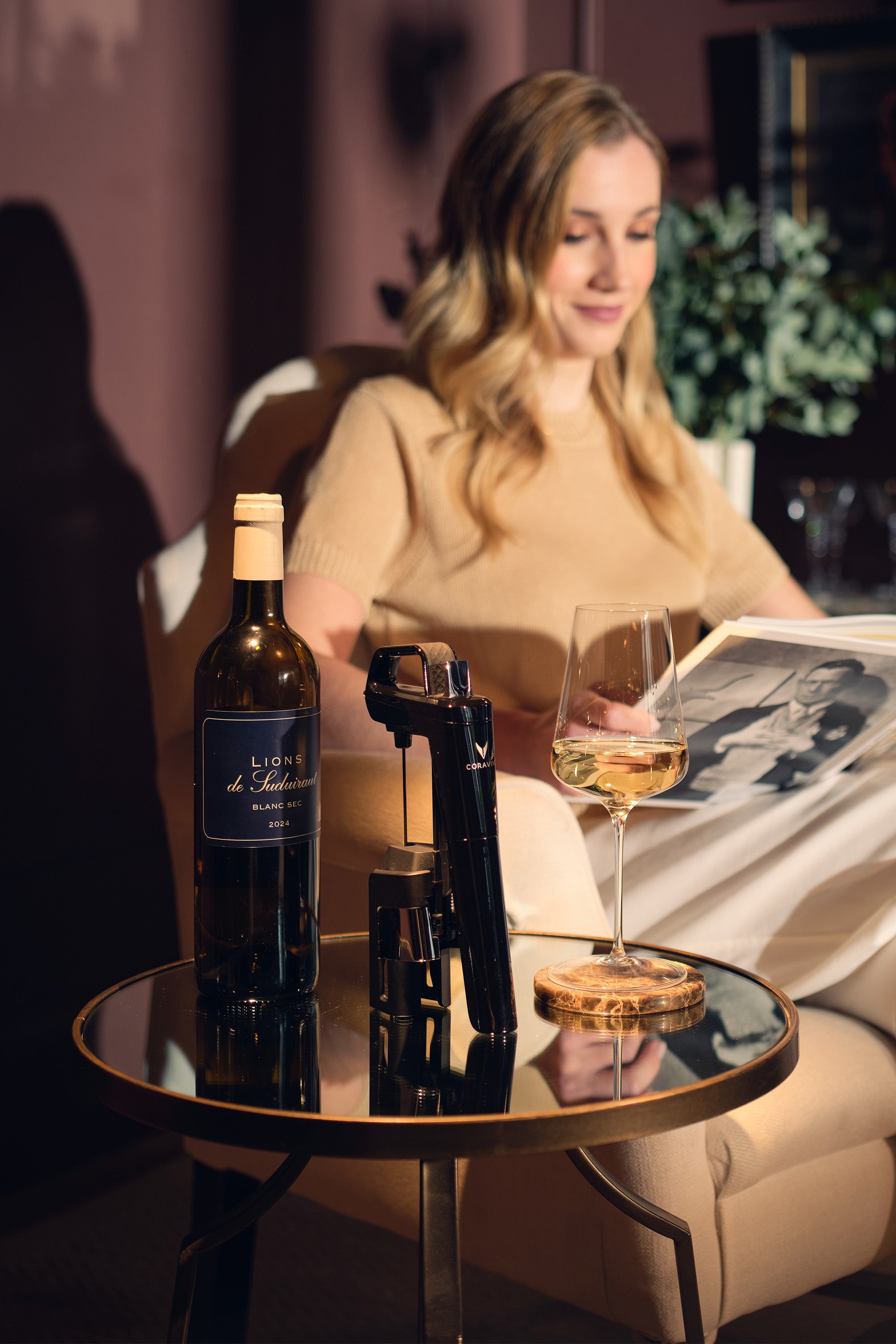 Woman sitting at a table with a bottle of wine, glass, and Coravin in a cozy indoor setting.
