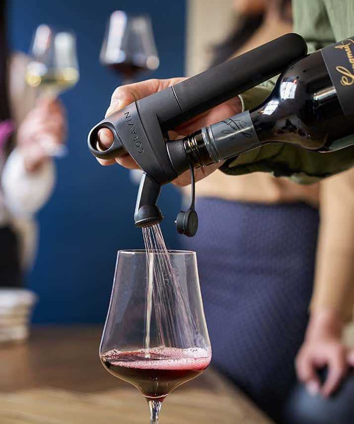 A hand uses a Coravin wine preservation system to pour red wine into a glass, with blurred attendees in the background.