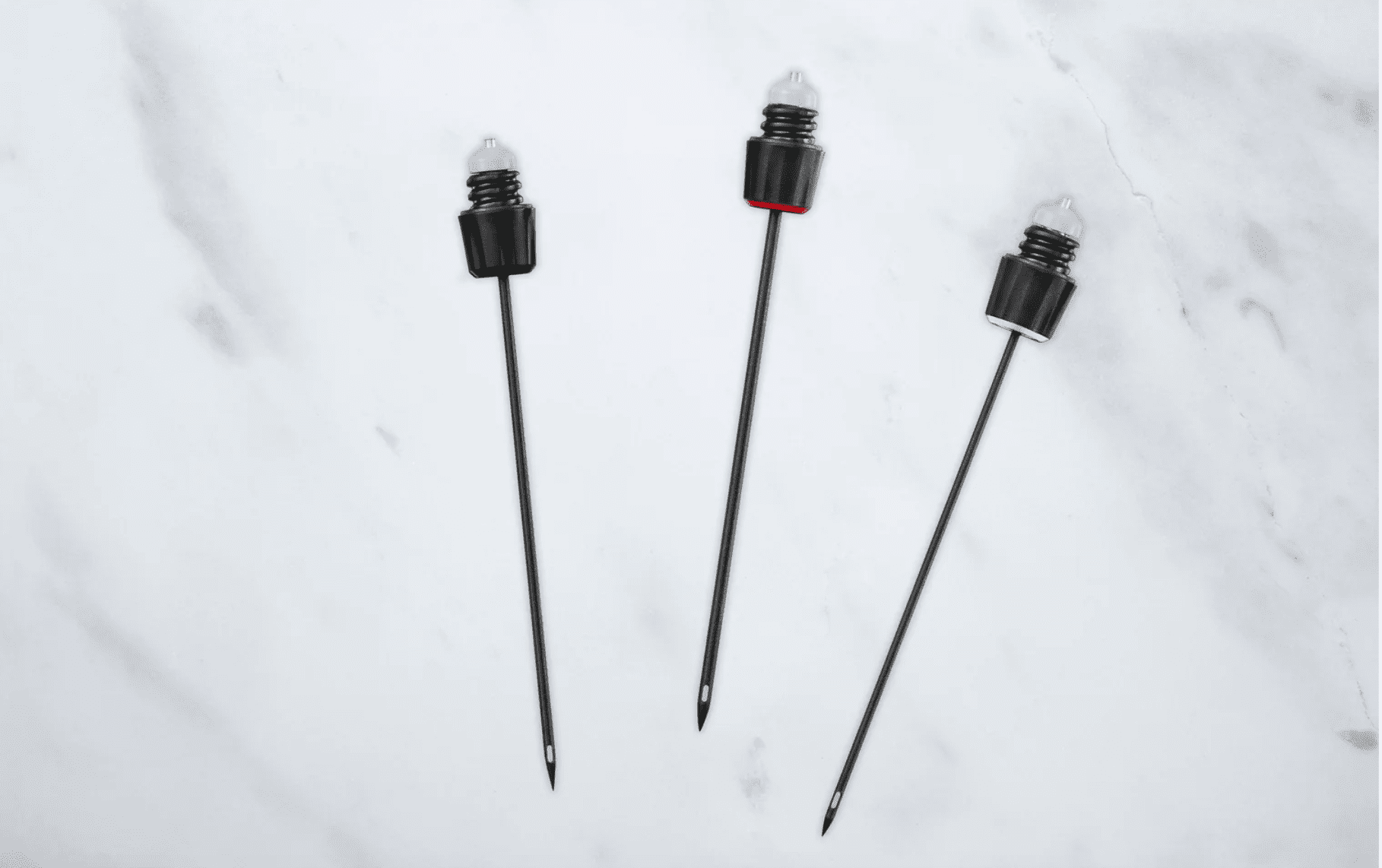 Three black barbecue skewers with pointed tips, one featuring a red accent near the handle, on a marble surface.